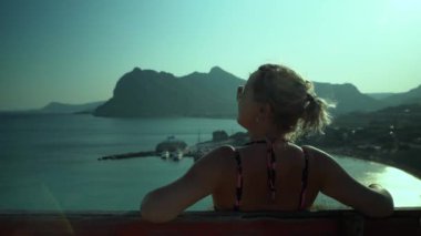 Rodos Adası - Yunanistan: Gün batımı / gün doğumunda kayalık tepeli bir Yunan manzarası / deniz manzarasına bakan bir bankta oturan beyaz bir genç kız / kadın. Doğanın tadını çıkaran kadın