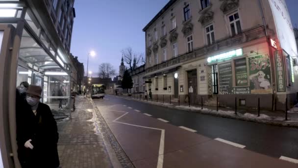 2021年2月27日 波兰克拉科夫 一名戴口罩 冬衣和帽子的女人站在公共汽车站台上 等待一辆公共汽车在大流行期间撞上擦亮的大楼 新的正常 欧洲公交车站和乘客等候 — 图库视频影像