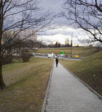 Krakow, Poland - March 12 , 2018: A footpath in the city with and trees with shredded leafless trees on both side and grass field against busy traffic on road during cloudy sky.