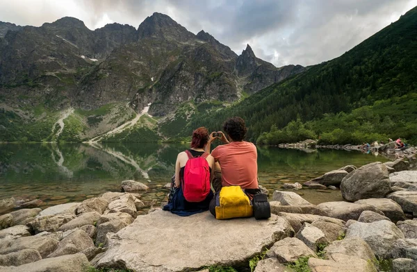 夏天在山上远足 一对年轻的女孩和一个男人坐在湖边休息 带着背包 健身探险和健康的生活方式 为山湖美景拍照 图库图片