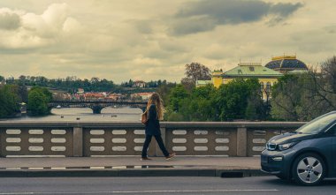 Prag Köprüsü manzaralı kişi yürüyüşü, Vltava Nehri, tarihi binalar, sarı barok mimari, bulutlu gökyüzü, şehir sokak manzarası, nehir kenarı