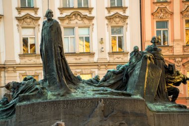 Prag meydanında yer alan büyük bir bronz heykel grubu, merkezi bir ayakta figürün ve yazılı tabanı ile çevreleyen figürlerin yer aldığı bir meydanda duruyor. Süslü pastel binalar tarihi bir kentsel zemin oluşturuyor..