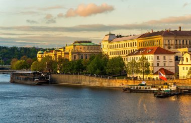 Prag nehir kenarının tarihi binaları, Vltava Nehri ve yolcu tekneleri, sıcak gün batımı ışığı ve bulutlu gökyüzü, ilkbahar ağaçları ve Çek Cumhuriyeti 'ndeki liman gezisi ile altın saat manzarası.
