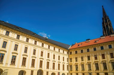 Prag Kalesi avlusunda mavi gökyüzünün altında Gotik katedrali olan barok sarı saray binaları var.