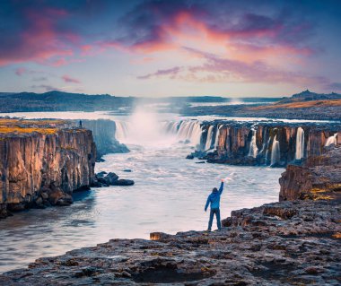 Jokulsa 'nın kıyısında ayakta duran bir turist el kaldırarak Fjollum Nehri' nin tadını çıkarıyor. Jokulsargljufur Ulusal Parkı, İzlanda 'daki Selfoss Şelalesinde renkli bir yaz sahnesi.