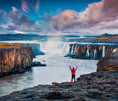 Jokulsa 'nın kıyısında ayakta duran turist ellerini kaldırarak gündoğumunun tadını çıkarıyor. Jokulsargljufur Ulusal Parkı, İzlanda 'daki Selfoss Şelalesinde renkli bir yaz sahnesi.