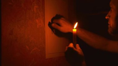 Elektrik kesintisi, elektrik yok. Karanlıkta elinde mum olan bir adam elektrik kesintisi sırasında evdeki elektrik santralini inceliyor..