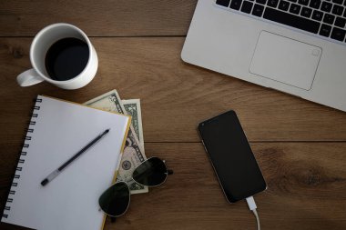 Üzerinde nesneler olan ahşap masa manzarası. Laptop, cep telefonu, not defteri, kahve fincanı, para, kalem ve güneş gözlüğü. Boşluğu kopyala.
