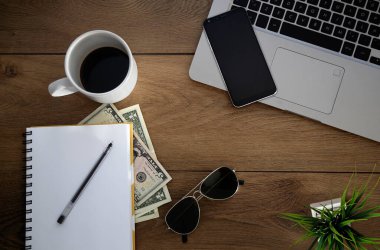 Üzerinde nesneler olan ahşap masa manzarası. Dizüstü bilgisayar, defter, kahve fincanı, para, bitki, kalem ve güneş gözlüğü. Boşluğu kopyala.