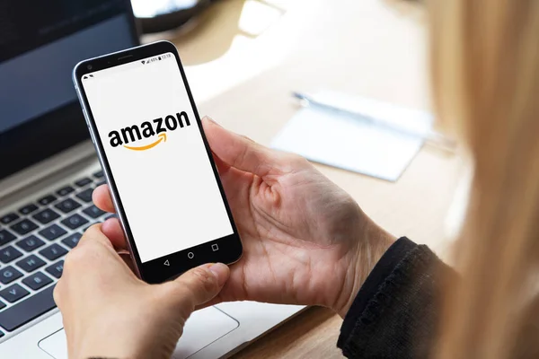 ROSARIO, ARGENTINA - 9 Kasım 2020: cep telefonu ekranında Amazon logosu. İş yerindeki sarışın kadın, dizüstü bilgisayarının önünde elinde akıllı telefonuyla..