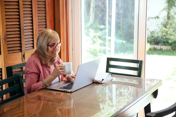 Senior woman with a laptop and coffee cup in a hand. Smiling grandmother in a wooden table with a silver computer.