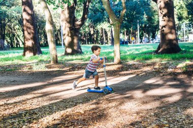 Parkta scooter kullanan bir çocuk. Çocuklar paten kaymayı öğreniyor. Küçük çocuk güneşli yaz gününde paten kayıyor. Güvenli sokaktaki çocuklar için açık hava aktivitesi. Anaokulu çocuğu için aktif spor.