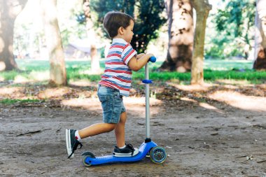 Parkta scooter kullanan bir çocuk. Çocuklar paten kaymayı öğreniyor. Küçük çocuk güneşli yaz gününde paten kayıyor. Güvenli sokaktaki çocuklar için açık hava aktivitesi. Anaokulu çocuğu için aktif spor.