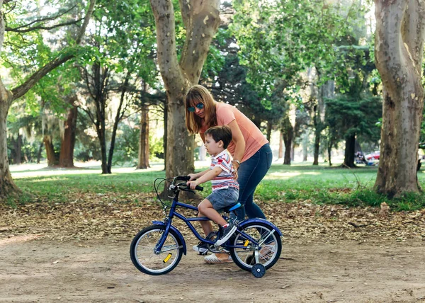 Mom teach to her little son to ride a bicycle. Small boy riding a blue bike in a dirty road in a forest park and helped by his mother. Bicycle with the training wheels.
