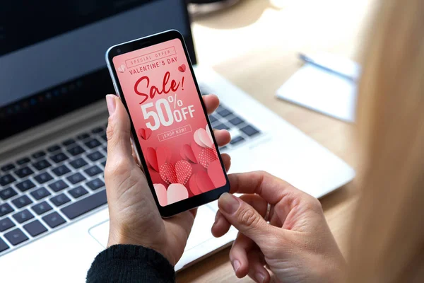 Sevgililer Günü 'nde cep telefonu ekranında indirim var. 14 Şubat 'ta indirimde olan akıllı telefonu elinde tutan genç bir kadın.% 50 indirimli. Hemen alışveriş yap..