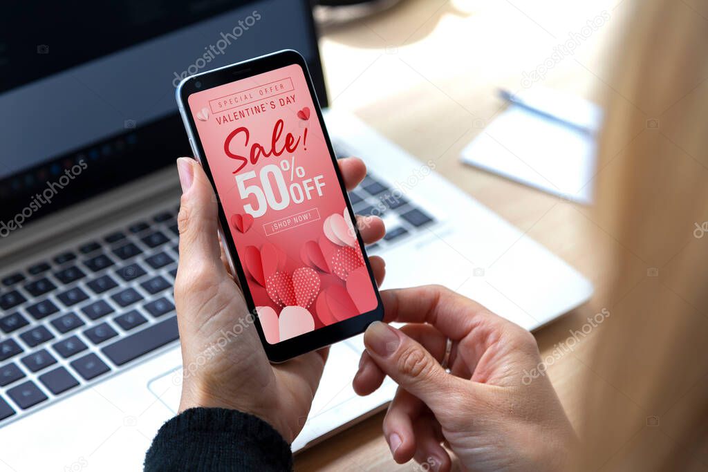 Valentine's Day offer sale in the screen of mobile phone. Young woman holding a smartphone with advertising on february 14 Special discount 50% off. Shop now.