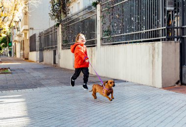 Mutlu küçük çocuk, köpeğiyle şehir kaldırımında koşuyor..
