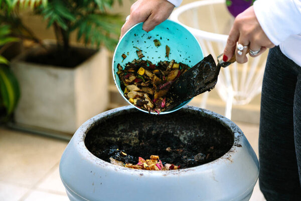 Woman doing compost in the backyard of his house. She is throwing waste of vegetables in the composter.