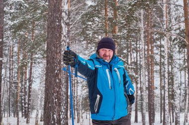 Mavi ceketli, siyah örgü şapkalı, kışın karlı bir ormanın arka planında kayak yapan mutlu yaşlı bir adamın portresi. Yakın plan.