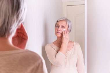 Kısa gri saçlı, sıradan giyimli, yaşlı, beyaz bir kadın, yüz derisini önemser..