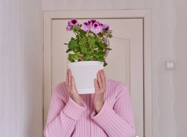 Elinde pelargonyum saksısı olan pembe hırkalı, kısa gri saçlı, yaşlı, beyaz bir kadın..