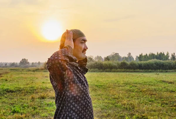 A Muslim senior man wearing a skullcap and traditional clothes prays at sunset