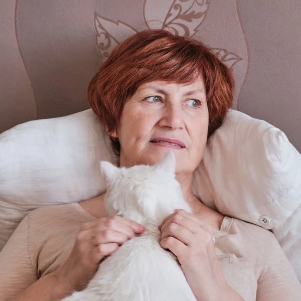 Idősebb nő ül, simogat egy aranyos fehér macskát, figyelmesen félrenéz. — Stock Fotó