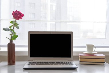 Kil şişesinde pembe gül, dizüstü bilgisayar, kitaplar ve kahve fincanı pencerenin yanındaki beyaz masada..