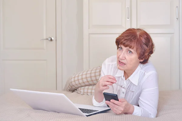 Senior woman wearing glasses, using mobile phone, laptop on the bed at home. — Stock Photo, Image