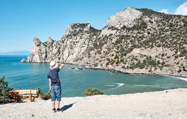 A boy in casual clothes standing on the shore of the bay and looking at the rocks. — Stock Photo, Image