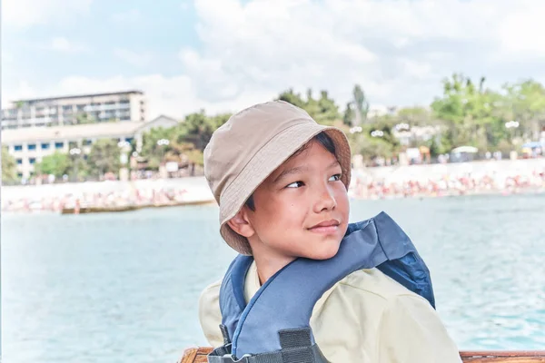 Un chico asiático en chaleco salvavidas flotando en barco de recreo en el mar y sonriendo. — Foto de Stock