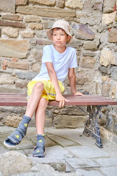 Cute Asian boy in casual summer clothes sitting on bench over background of stone ancient wall. — Zdjęcie stockowe
