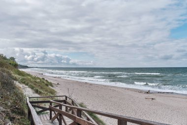 Dalgalı güzel deniz manzarası, kumlu plaj, eski dalgalı sular, bulutlu gökyüzü ve ön planda ahşap merdivenler. Baltık Denizi, Curonian Tükürüğü.