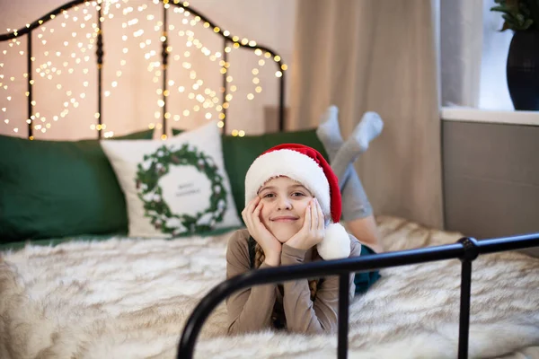 サンタクロースの帽子のかわいい女の子の笑顔は お祝いの新年やクリスマスの雰囲気の中でベッドの上に横たわっています クリスマスライト付きの魔法のボケ メリークリスマスとハッピーニューイヤーのコンセプト 選択的焦点 — ストック写真