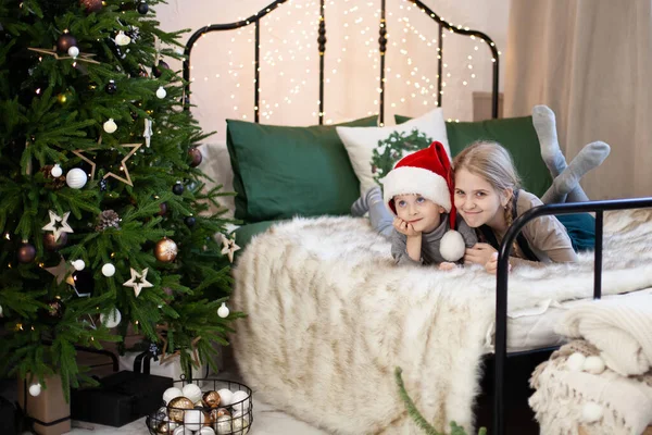 サンタクロースの帽子のかわいい女の子と男の子の笑顔は クリスマスツリーの近くのベッドの上に横たわっています クリスマスライト付きの魔法のボケ メリークリスマスとハッピーニューイヤーのコンセプト 選択的焦点 — ストック写真
