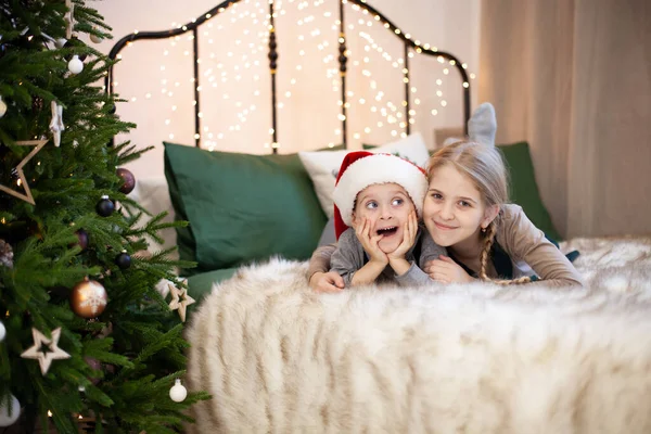 サンタクロースの帽子のかわいい女の子と感情的な男の子は クリスマスツリーの近くのベッドの上に横たわっています クリスマスライト付きの魔法のボケ 子供たちはメリークリスマスとハッピーニューイヤーを待ちます 選択的焦点 — ストック写真