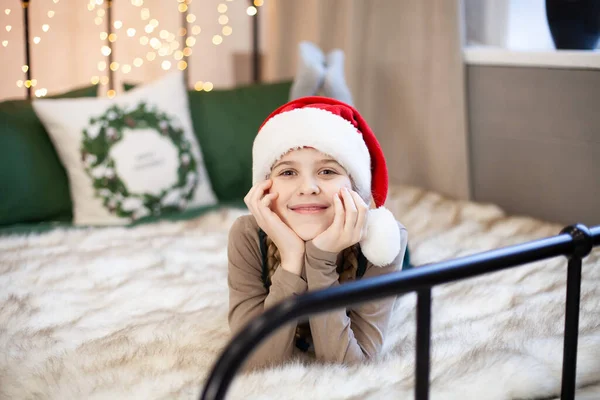 サンタクロースの帽子のかわいい女の子の笑顔は お祝いの新年やクリスマスの雰囲気の中でベッドの上に横たわっています クリスマスライト付きの魔法のボケ メリークリスマスとハッピーニューイヤーのコンセプト 選択的焦点 — ストック写真