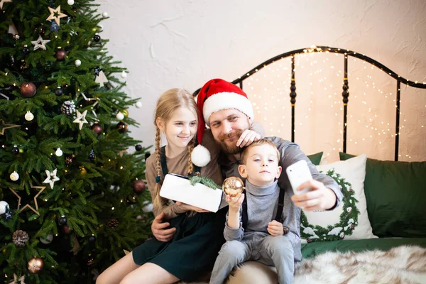サンタクロースの帽子を被った父親と彼の2人の子供は スマートフォンを使用してオンラインで家族を祝福したり ギフトボックスを一緒に自撮りしたりします 大流行中のクリスマスオンラインお祝い — ストック写真