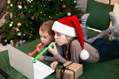 Noel Baba şapkası takan çocuklar arkadaşlarını ve akrabalarını parti boynuzlarını kullanarak kutluyorlar. Noel online iletişimi ve salgın sırasında tebrikler.. 