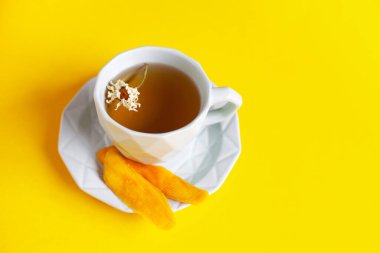 Sarı arka planda bir fincan bitkisel papatya çayı ve kurutulmuş mango dilimleri. Sağlıklı kafeinsiz içecek ve doğal kurutulmuş meyve kavramı. Vejetaryen tatlısı, çay molası. Seçici odak.