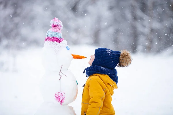 冬の雪の日に雪だるまの鼻を見る明るいニットの服を着た少年 愛の冬 明るい色とアクティブな屋外レジャーコンセプト — ストック写真
