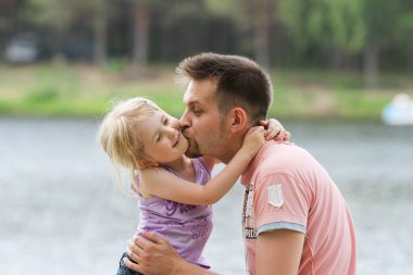 Babası gölün kıyısında küçük tatlı kızını kucaklıyor ve öpüyor. Aile, ülkede birlikte vakit geçiriyor. Baba-kız ilişkisi, babalar günü, aile bakımı. Seçici yumuşak odak.