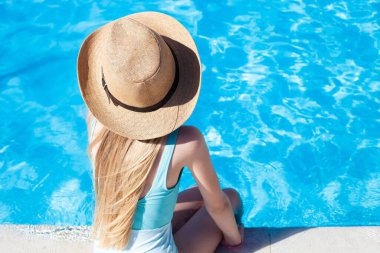 Mavi mayo ve bej hasır şapka giyen sarışın kız havuz kenarında oturuyor. Arka plan, boşluğu kopyala. Seyahat, tatil, dinlenme, yaz havası..