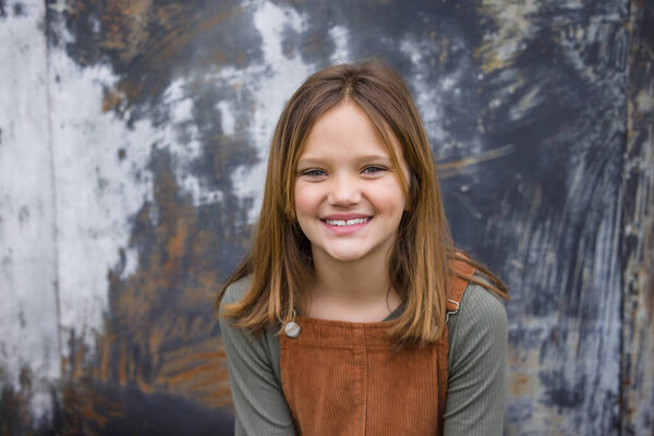 Young girl in a brown overall dress in the winter sitting near an urban warehouse wall