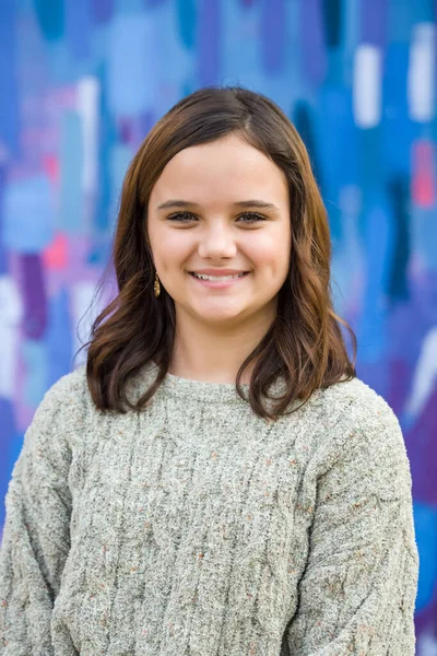 Junges Mädchen, das im Winter einen Pullover trägt, steht draußen neben einer hellen Wand mit blauen, rosa und lila. — Stockfoto