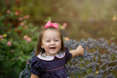 Donanma elbisesi ve pembe kurdeleli küçük bir kız demir bir bankta oturuyor.