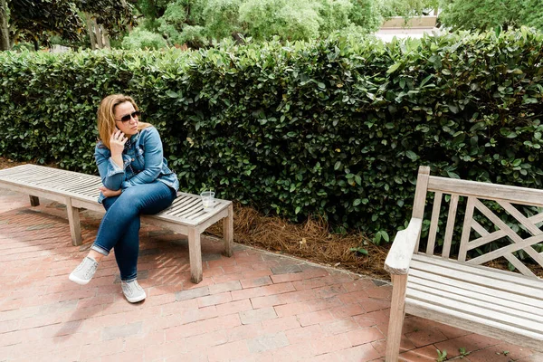 Kobieta w okularach przeciwsłonecznych siedzi na ławce i rozmawia przez telefon. — Zdjęcie stockowe