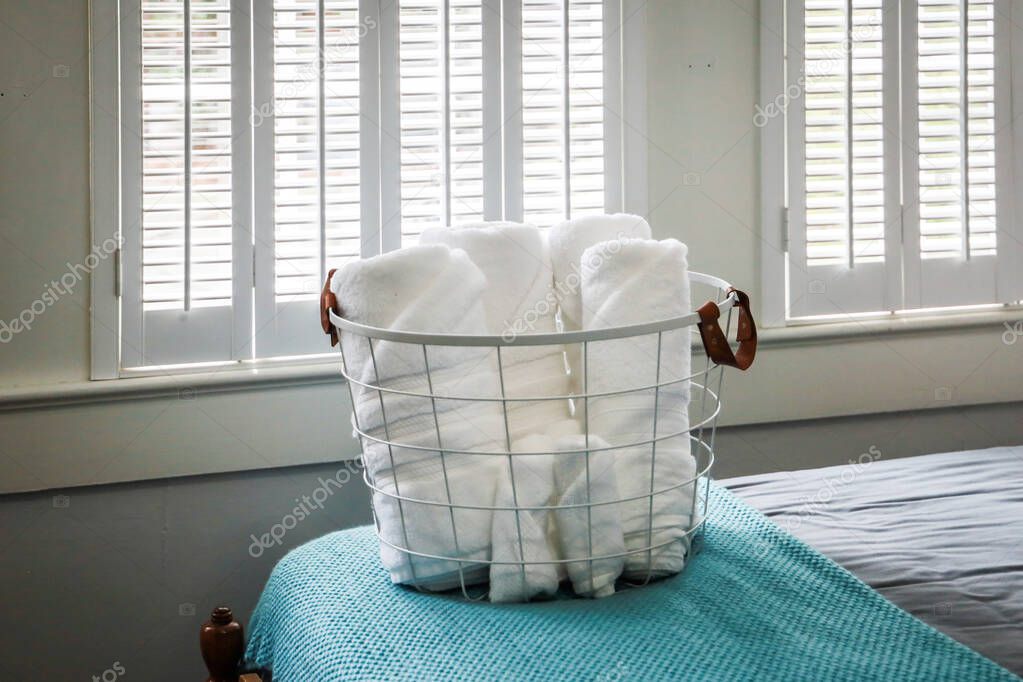 A basket of organized clean rolled white towels for guests sitting on a bed in a guest bedroom