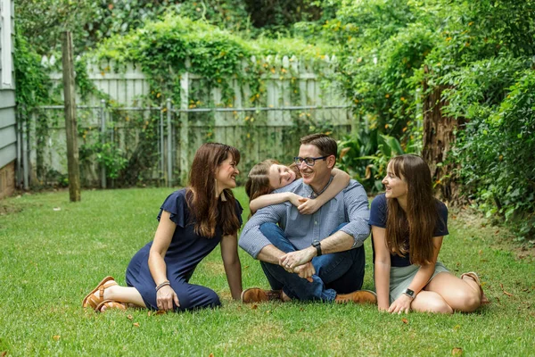 A family of a mother and father and two daughters sitting outside in the grass of their backyard witht the youngest daughter hugging dad from behind