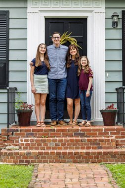 Bir anne, baba ve iki kızı olan bir aile küçük mavi bir yazlık evin tuğla verandasında duruyorlar.